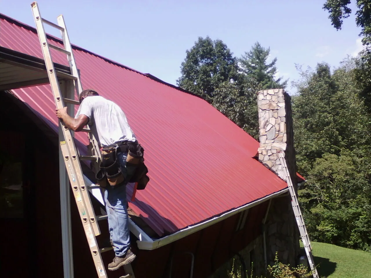 Roofer on a ladder beside a red metal roof and stone chimney — Soffit & Fascia Repair by Roofing Brothers in Severna Park